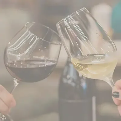 Zwei Personen stossen mit einem Rotwein- und einem Weissweinglas an, im Hintergrund steht eine Weinflasche.